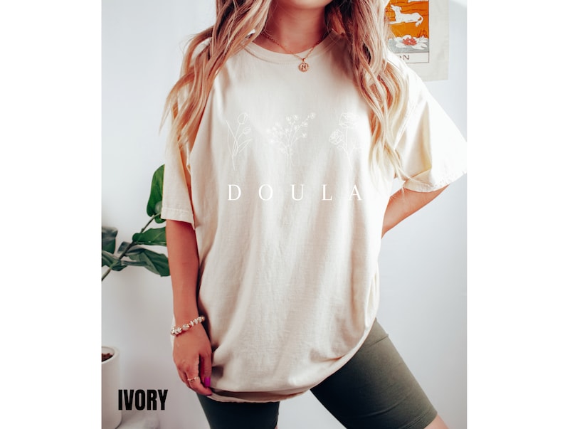 May include: A cream colored t-shirt with a white floral design and the word "DOULA" printed on the front. The t-shirt is being worn by a person with long blonde hair. The word "IVORY" is printed in black at the bottom of the image.