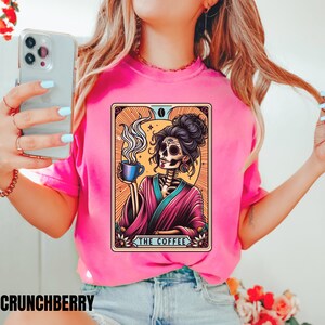 May include: A pink t-shirt with a graphic of a skeleton woman holding a cup of coffee. The graphic is in a tarot card style with the text "The Coffee" at the bottom.