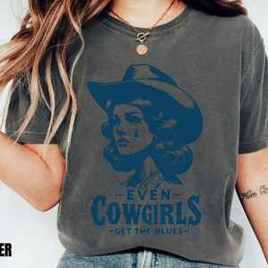 May include: A dark gray t-shirt with a blue graphic of a woman wearing a cowboy hat and the text "Even Cowgirls Get The Blues".