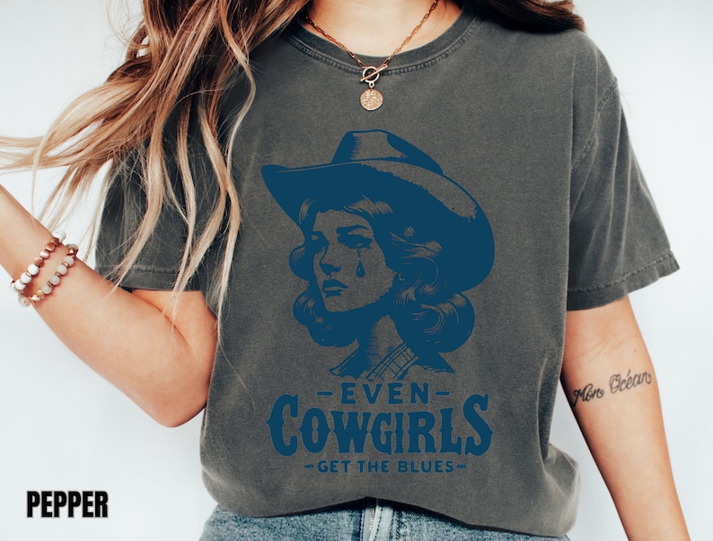 May include: A dark gray t-shirt with a blue graphic of a woman wearing a cowboy hat and the text "Even Cowgirls Get The Blues".
