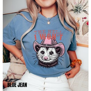 Pu&ograve; includere: Una maglietta blu con una grafica di un opossum che indossa un cappello da cowboy rosa. Il testo "THE HELL I WON'T" &egrave; stampato sulla maglietta.
