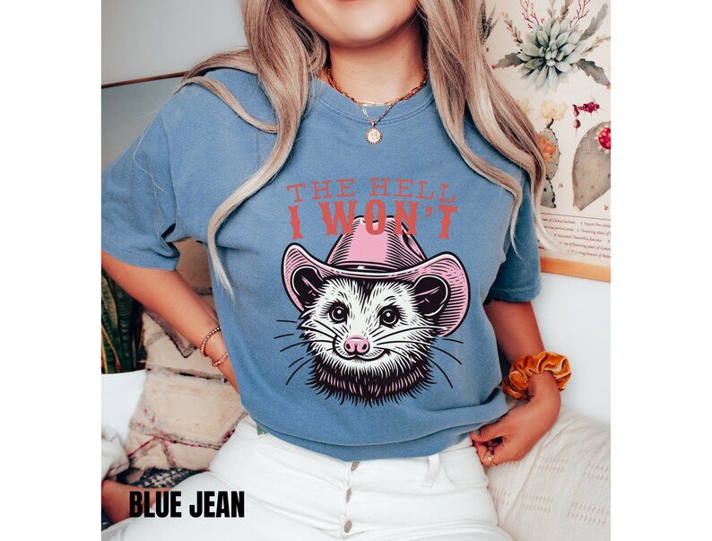 Pu&ograve; includere: Una maglietta blu con una grafica di un opossum che indossa un cappello da cowboy rosa. Il testo "THE HELL I WON'T" &egrave; stampato sulla maglietta.