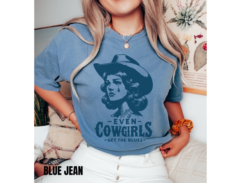 May include: A blue jean t-shirt with a graphic of a woman wearing a cowboy hat and a plaid shirt. The text on the shirt reads "Even Cowgirls Get The Blues".