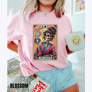 May include: A pink t-shirt with a graphic of a skeleton holding a cup of coffee. The graphic is in a tarot card style with the text "The Coffee" at the bottom.