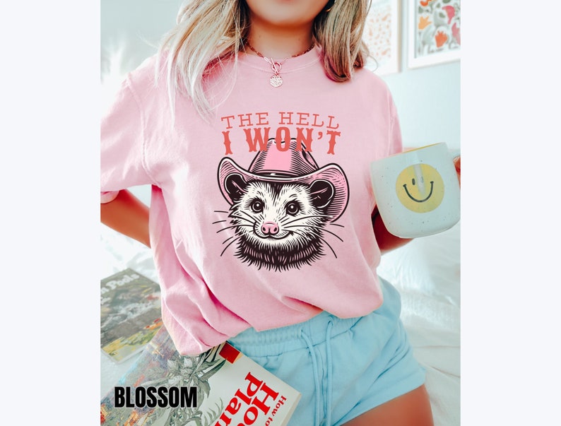 Pu&ograve; includere: Maglietta rosa con un'immagine di un opossum che indossa un cappello da cowboy rosa e il testo "The Hell I Won't".