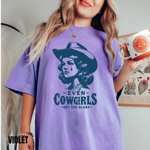 May include: A purple t-shirt with a graphic of a woman wearing a cowboy hat and the text "Even Cowgirls Get the Blues".