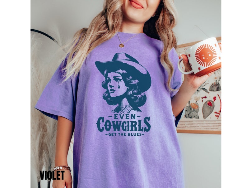 May include: A purple t-shirt with a graphic of a woman wearing a cowboy hat and the text "Even Cowgirls Get the Blues".