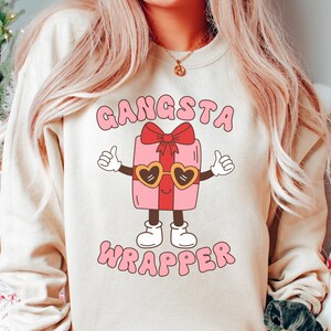 Puede incluir: Una sudadera de color crema con el texto "Gangsta Wrapper" en letras rosas y rojas. Una caja de regalo rosa de dibujos animados con un lazo rojo y gafas de sol está en el centro del texto.