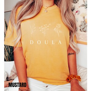 May include: A mustard yellow t-shirt with the word "DOULA" printed in white. The shirt features a simple line drawing of three flowers.