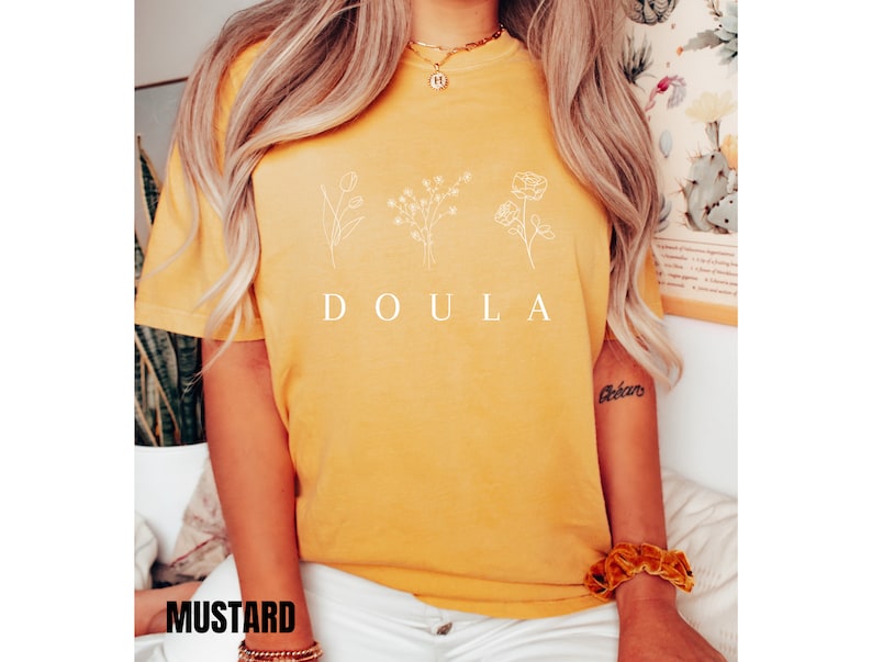 May include: A mustard yellow t-shirt with the word "DOULA" printed in white. The shirt features a simple line drawing of three flowers.