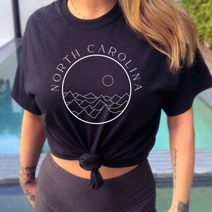 K&ouml;nnte beinhalten: Schwarzes T-Shirt mit einem wei&szlig;en Kreis-Motiv mit dem Text "NORTH CAROLINA" und einer stilisierten Bergkette mit einer Sonne.