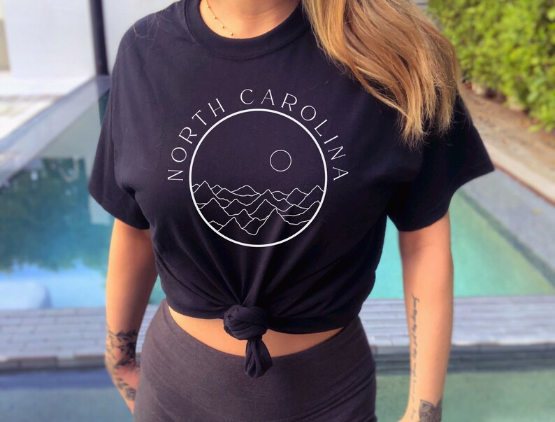 K&ouml;nnte beinhalten: Schwarzes T-Shirt mit einem wei&szlig;en Kreis-Motiv mit dem Text "NORTH CAROLINA" und einer stilisierten Bergkette mit einer Sonne.