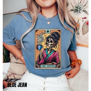 May include: A blue t-shirt with a graphic of a skeleton woman holding a cup of coffee. The graphic is in a tarot card style with the text "The Coffee" at the bottom.