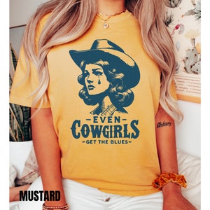 May include: A mustard yellow t-shirt with a vintage illustration of a woman wearing a cowboy hat and the text "Even Cowgirls Get The Blues".