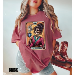 May include: A maroon t-shirt with a graphic of a skeleton woman holding a cup of coffee. The graphic is in a tarot card style with the text "The Coffee" at the bottom.