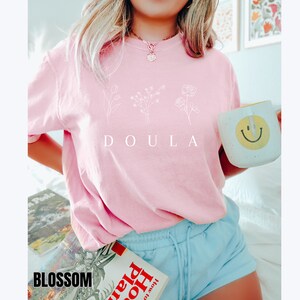 May include: A pink t-shirt with a white graphic of three flowers and the word "DOULA" printed on the front.