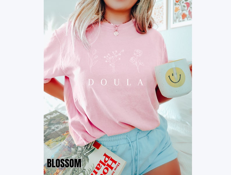 May include: A pink t-shirt with a white graphic of three flowers and the word "DOULA" printed on the front.