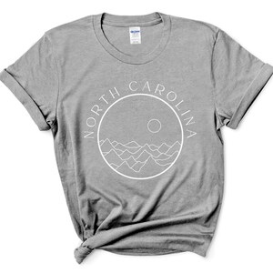 K&ouml;nnte beinhalten: Ein heidefarbenes T-Shirt mit einer wei&szlig;en Grafik eines Kreises mit den Worten "NORTH CAROLINA" um den Au&szlig;enrand und einer Sonne und Bergen innerhalb des Kreises.