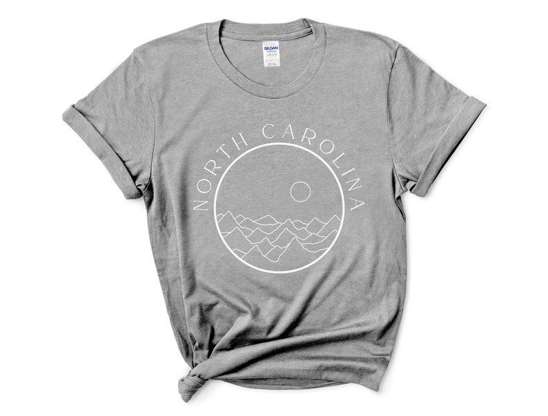 K&ouml;nnte beinhalten: Ein heidefarbenes T-Shirt mit einer wei&szlig;en Grafik eines Kreises mit den Worten "NORTH CAROLINA" um den Au&szlig;enrand und einer Sonne und Bergen innerhalb des Kreises.