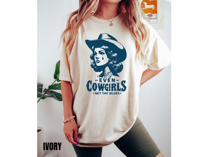 May include: A cream colored t-shirt with a blue graphic of a woman wearing a cowboy hat and the text "Even Cowgirls Get The Blues".