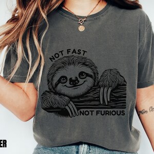 May include: A dark gray t-shirt with a black and white illustration of a sloth on a branch. The text "NOT FAST NOT FURIOUS" is printed on the shirt.