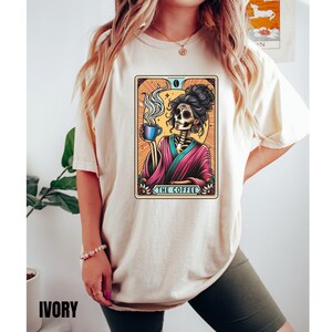 May include: A beige t-shirt with a colorful illustration of a skeleton holding a cup of coffee. The illustration is in the style of a tarot card and reads "The Coffee".