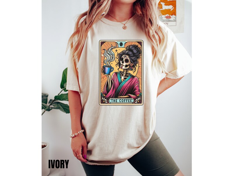 May include: A beige t-shirt with a colorful illustration of a skeleton holding a cup of coffee. The illustration is in the style of a tarot card and reads "The Coffee".