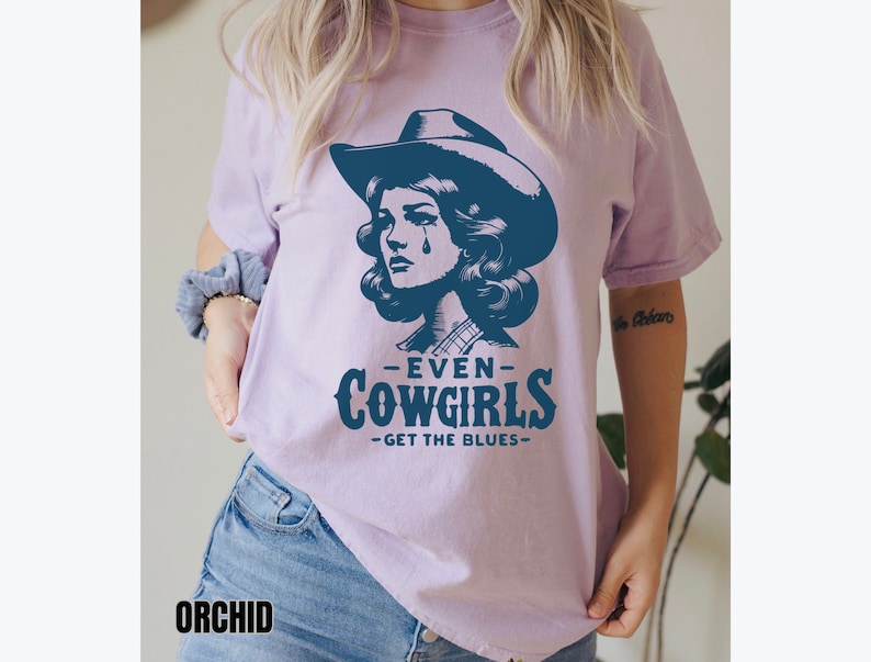 May include: A purple t-shirt with a graphic of a cowgirl wearing a hat and a teardrop on her cheek. The text on the shirt reads "Even Cowgirls Get The Blues".