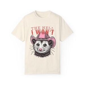 Pu&ograve; includere: Una t-shirt beige con una grafica di un opossum che indossa un cappello da cowboy rosa. Il testo "THE HELL I WON'T" &egrave; stampato in rosso sopra l'opossum.