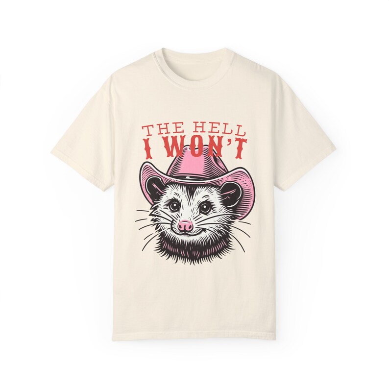 Pu&ograve; includere: Una t-shirt beige con una grafica di un opossum che indossa un cappello da cowboy rosa. Il testo "THE HELL I WON'T" &egrave; stampato in rosso sopra l'opossum.