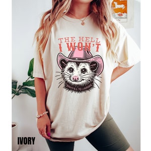 Pu&ograve; includere: Una t-shirt color crema con una grafica di un opossum che indossa un cappello da cowboy rosa. Il testo "The Hell I Won't" &egrave; stampato sulla t-shirt.