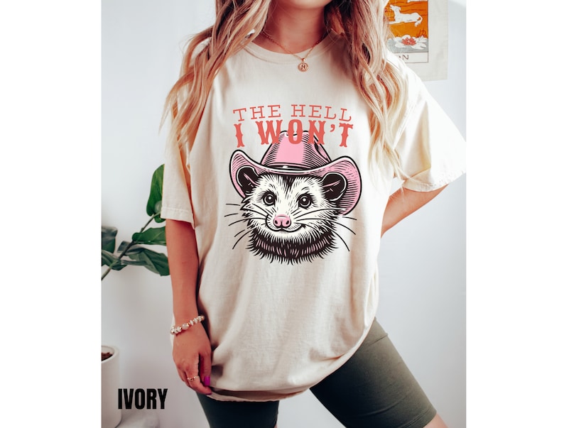 Pu&ograve; includere: Una t-shirt color crema con una grafica di un opossum che indossa un cappello da cowboy rosa. Il testo "The Hell I Won't" &egrave; stampato sulla t-shirt.