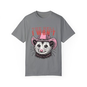 Pu&ograve; includere: Maglietta grigia con un opossum dei cartoni animati che indossa un cappello da cowboy rosa. Il testo "THE HELL I WON'T" &egrave; stampato sulla maglietta.