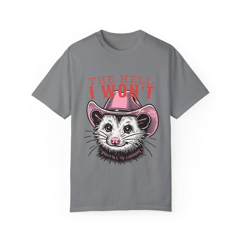 Pu&ograve; includere: Maglietta grigia con un opossum dei cartoni animati che indossa un cappello da cowboy rosa. Il testo "THE HELL I WON'T" &egrave; stampato sulla maglietta.