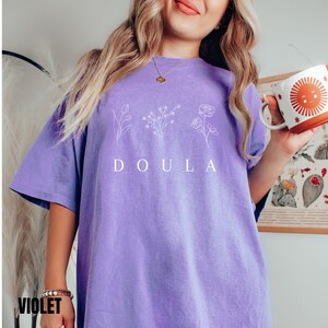 May include: A purple t-shirt with a white graphic of flowers and the word "DOULA" printed on the front.