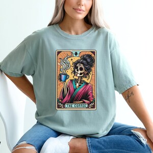 May include: A light blue t-shirt with a graphic of a skeleton woman holding a cup of coffee. The graphic is in a vintage tarot card style with the text "The Coffee" at the bottom.