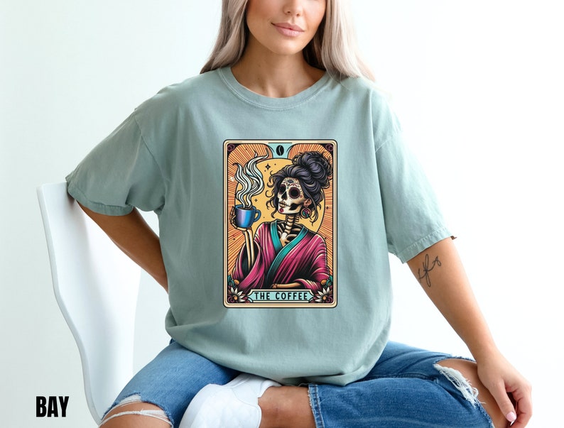 May include: A light blue t-shirt with a graphic of a skeleton woman holding a cup of coffee. The graphic is in a vintage tarot card style with the text "The Coffee" at the bottom.