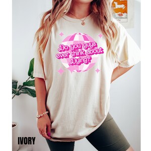 May include: A light beige t-shirt with a pink disco ball graphic and the text "Do you guys ever think about Dying?" printed on the front. The shirt is being modeled by a person wearing green shorts.