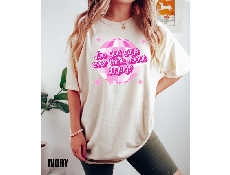 May include: A light beige t-shirt with a pink disco ball graphic and the text "Do you guys ever think about Dying?" printed on the front. The shirt is being modeled by a person wearing green shorts.