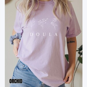 May include: A lavender t-shirt with a white graphic of three stylized flowers and the word "DOULA" printed on the front.