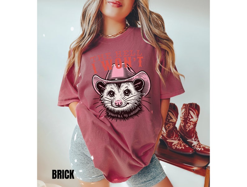 Pu&ograve; includere: Una maglietta color marsala con una grafica di un opossum che indossa un cappello da cowboy rosa. Il testo "The Hell I Won't" &egrave; stampato sulla maglietta.