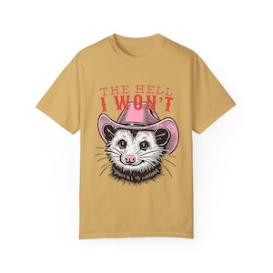 Pu&ograve; includere: Una maglietta color beige con un opossum dei cartoni animati che indossa un cappello da cowboy rosa. Il testo "THE HELL I WON'T" &egrave; stampato in rosso sulla maglietta.