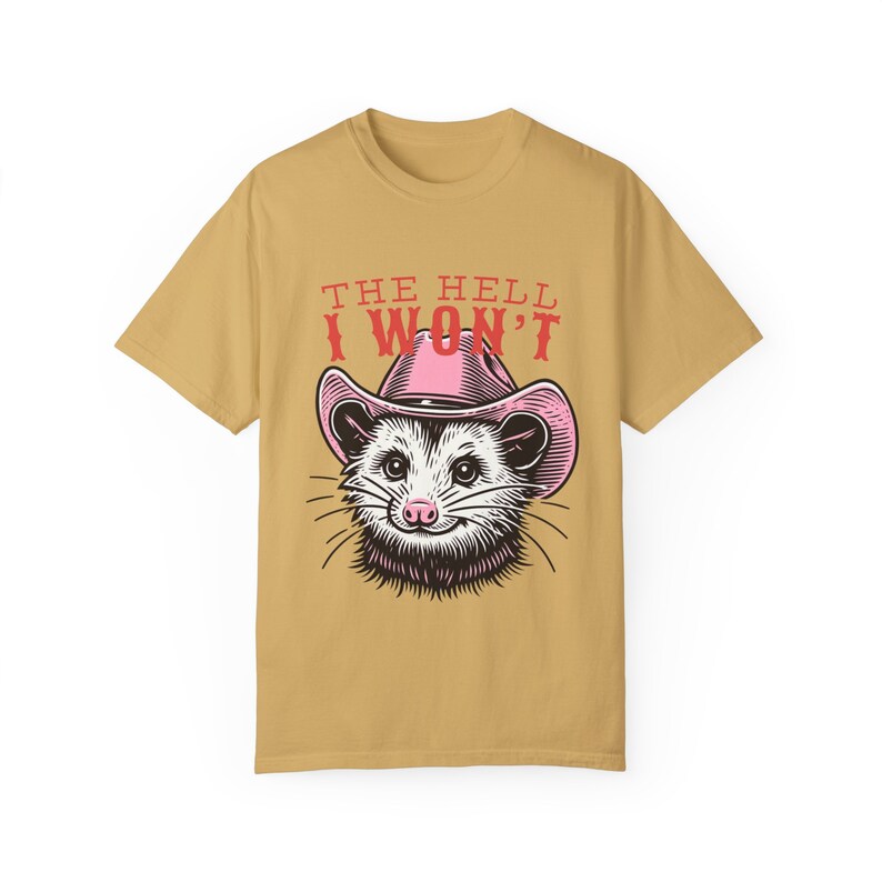 Pu&ograve; includere: Una maglietta color beige con un opossum dei cartoni animati che indossa un cappello da cowboy rosa. Il testo "THE HELL I WON'T" &egrave; stampato in rosso sulla maglietta.