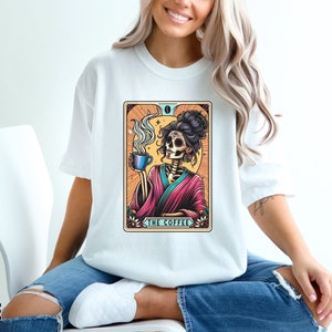 May include: White t-shirt with a colorful illustration of a skeleton woman holding a cup of coffee. The illustration is framed with a decorative border and the text "The Coffee" is printed below the image.