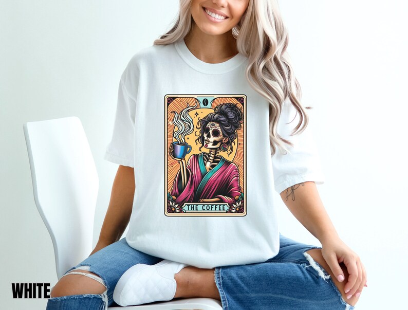 May include: White t-shirt with a colorful illustration of a skeleton woman holding a cup of coffee. The illustration is framed with a decorative border and the text "The Coffee" is printed below the image.
