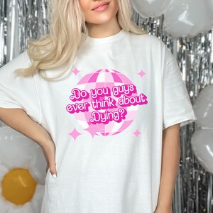 May include: White t-shirt with a pink disco ball graphic and the text "Do you guys ever think about Dying?"