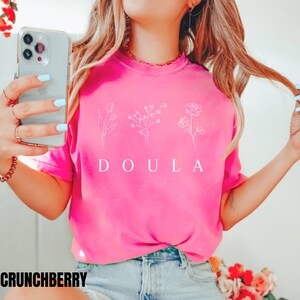 May include: A pink t-shirt with a white graphic of three flowers and the word "DOULA" printed on the front.