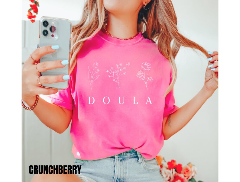 May include: A pink t-shirt with a white graphic of three flowers and the word "DOULA" printed on the front.