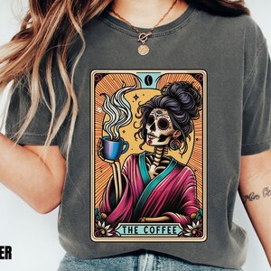 May include: A gray t-shirt with a colorful illustration of a skeleton woman holding a cup of coffee. The illustration is in the style of a tarot card and includes the text "The Coffee".