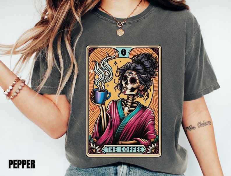 May include: A gray t-shirt with a colorful illustration of a skeleton woman holding a cup of coffee. The illustration is in the style of a tarot card and includes the text "The Coffee".